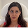A woman with long dark hair and red lipstick is seated, facing the camera in a red top against a light background, ready to share her reviews and testimonials.